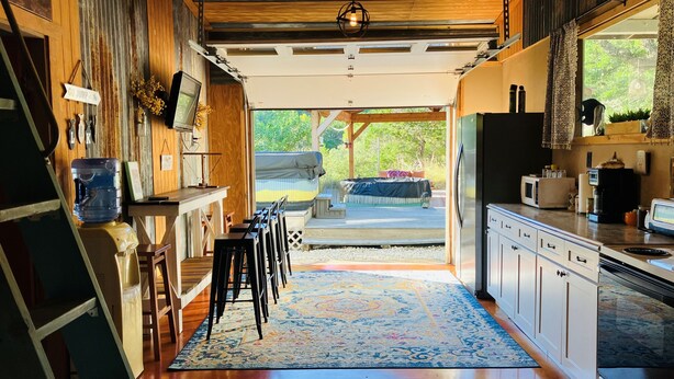 A spacious kitchen with a large island, wooden cabinetry, and a view to an outdoor deck. - Tiny cabin/guest house with hot tub and pool surrounded by nature (Lakehills)