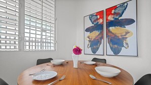 A dining area with a round wooden table, white plates, and silverware. There are two abstract paintings on the wall and a vase with flowers on the table.