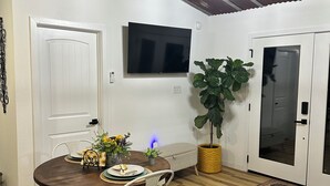Un comedor con una mesa de madera, sillas blancas, una planta en maceta y un televisor de pantalla plana montado en la pared.