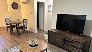 A living room with a round wooden coffee table, a television on a wooden stand, a dining area with a table and chairs, and a patterned rug. - The Relaxing House - Quiet 2 Bedroom Home (Raymond)