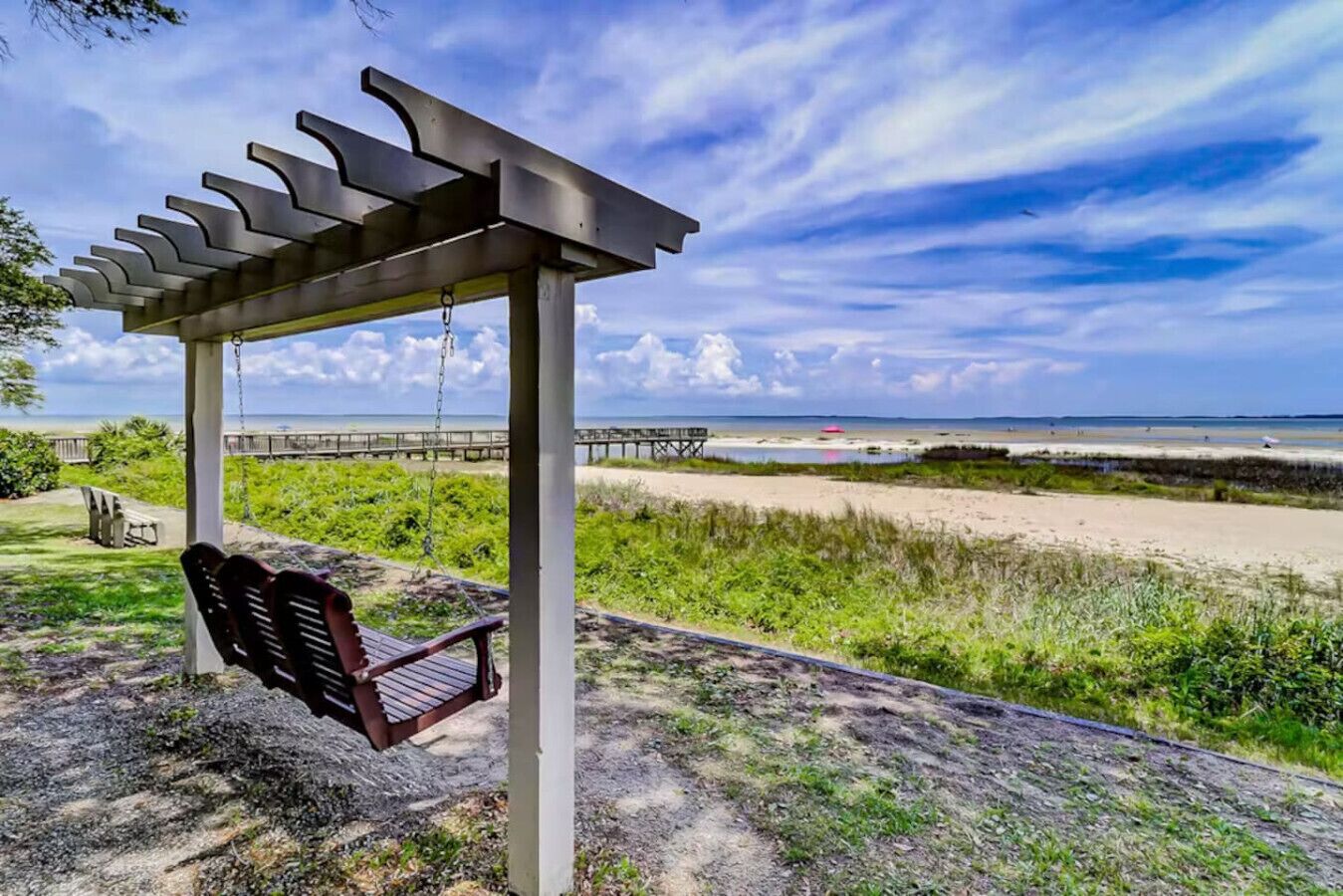 Ein überdachter Sitzbereich im Freien mit Schaukelstuhl, Holzbank und Blick auf Strand und Meer.