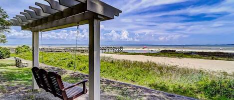 A covered outdoor seating area with a swing chair, a wooden bench, and a view of the beach and ocean.