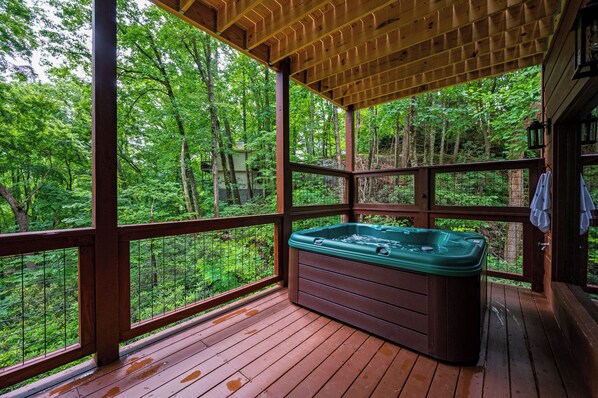 Une terrasse en bois dotée d’un jacuzzi, entourée d’une rambarde en bois et de panneaux de verre, et offrant une vue sur une zone boisée.