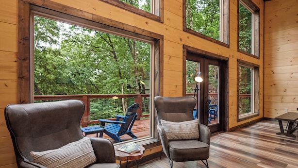 A wooden cabin with two leather chairs, a small table, and a large window overlooking a deck.