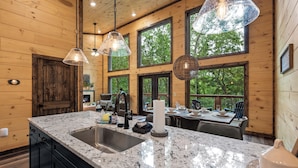A kitchen with granite countertops, a stainless steel sink, and wooden cabinetry. There are large windows providing natural light and views of the outdoors.