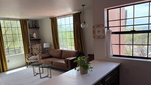 Una sala de estar acogedora con un sofá, una mesita y una planta en maceta. Cuenta con ventanas grandes con cortinas, una lámpara de techo y vistas al exterior con vegetación.