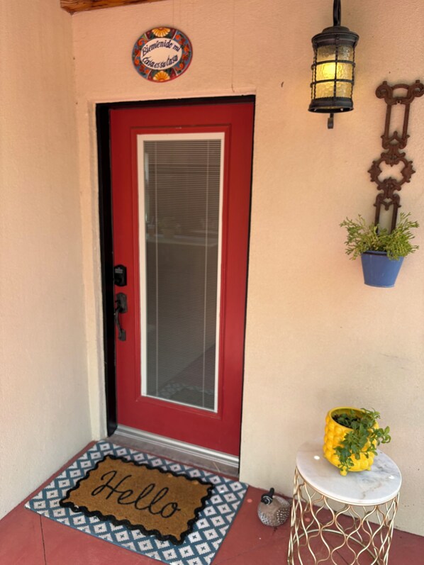 Una puerta roja con un panel de vidrio, un felpudo que dice 'Hello', un farol y una planta en maceta sobre una mesita.