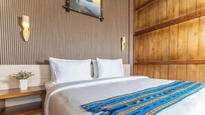 Una cama bien hecha con sábanas blancas y una colcha a rayas azul y amarilla. Tiene un cabecero de madera y una pequeña maceta con una planta en la mesita de noche.