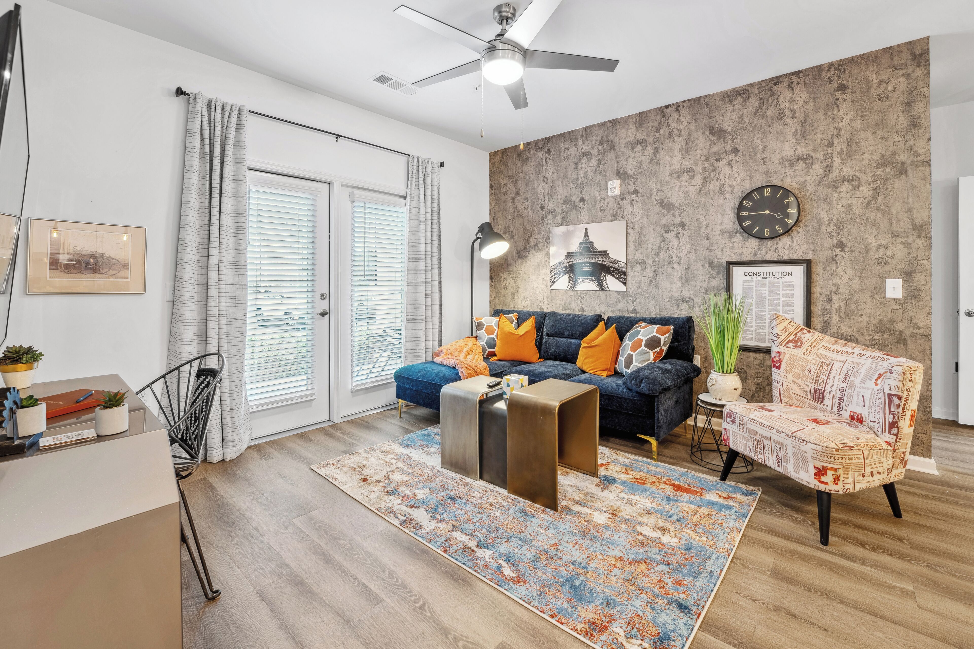 A modern living room with a dark blue sofa, a patterned rug, a ceiling fan, and a wall clock.