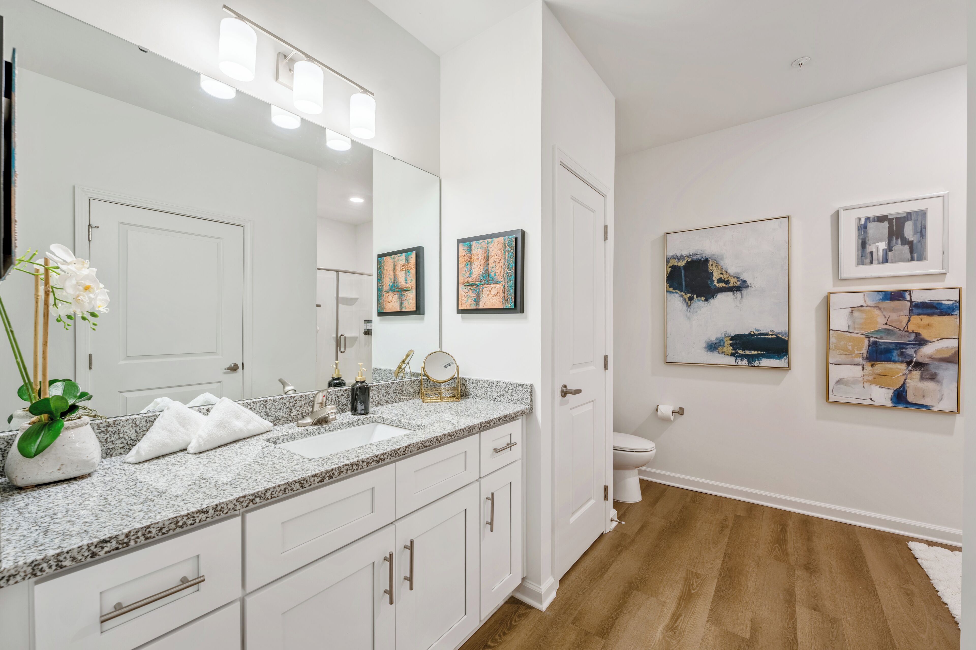 Een moderne badkamer met een granieten aanrechtblad, witte kastjes, een grote spiegel en kunstwerken aan de muur.