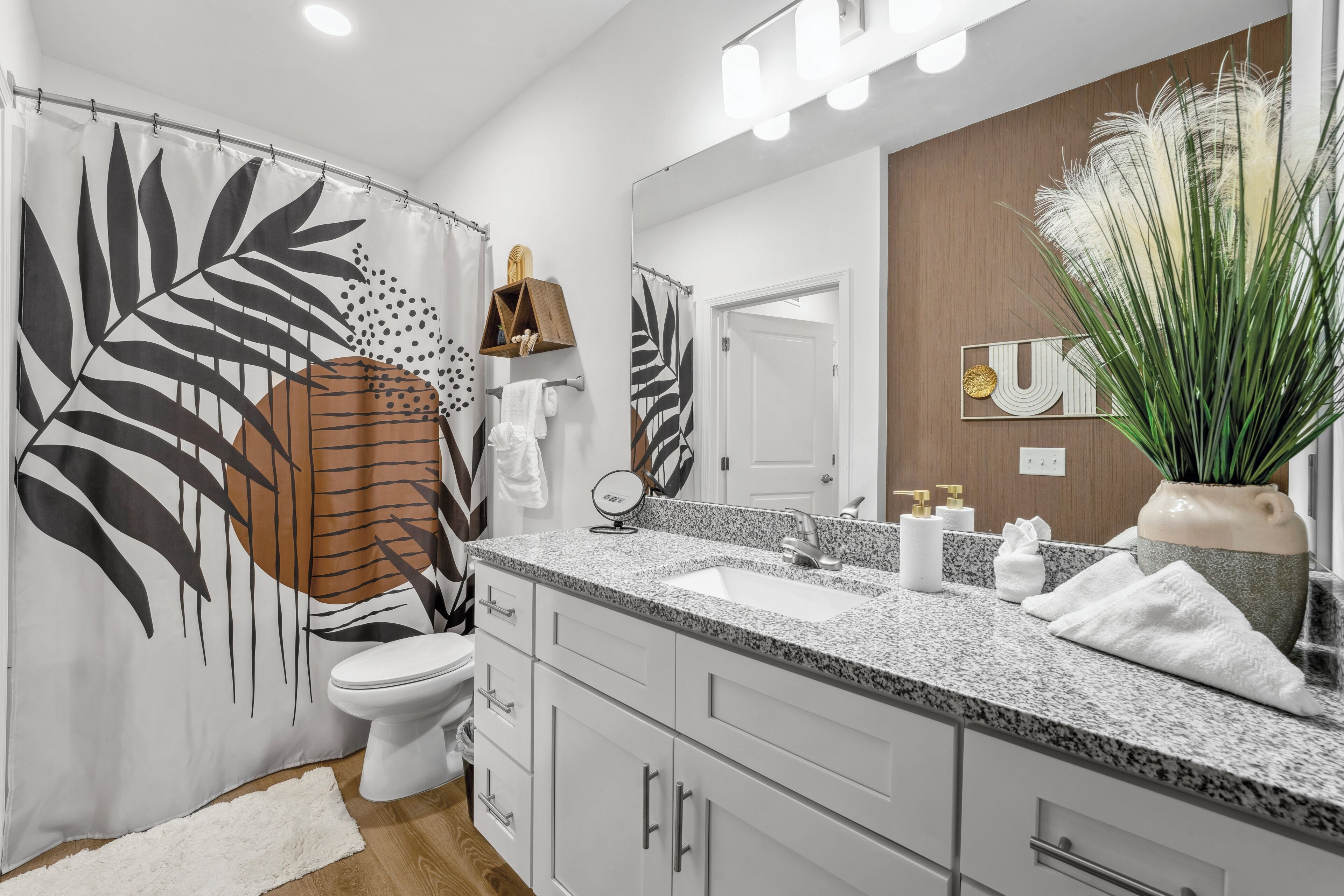 A modern bathroom with a shower curtain featuring a leaf pattern, a white toilet, a granite countertop sink, and a large mirror.