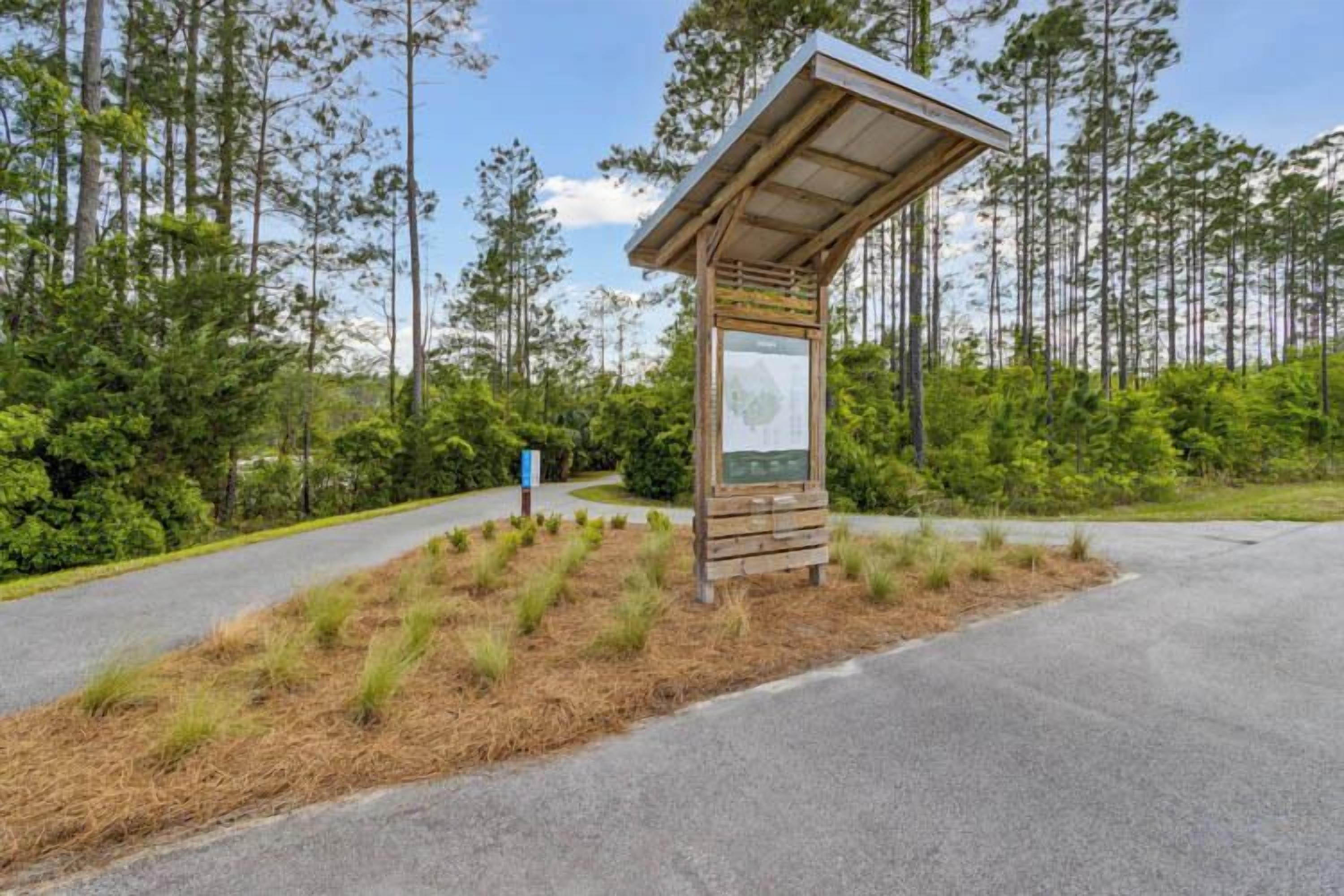 A wooden information kiosk with a map, surrounded by a landscaped area with pine trees and a paved road.