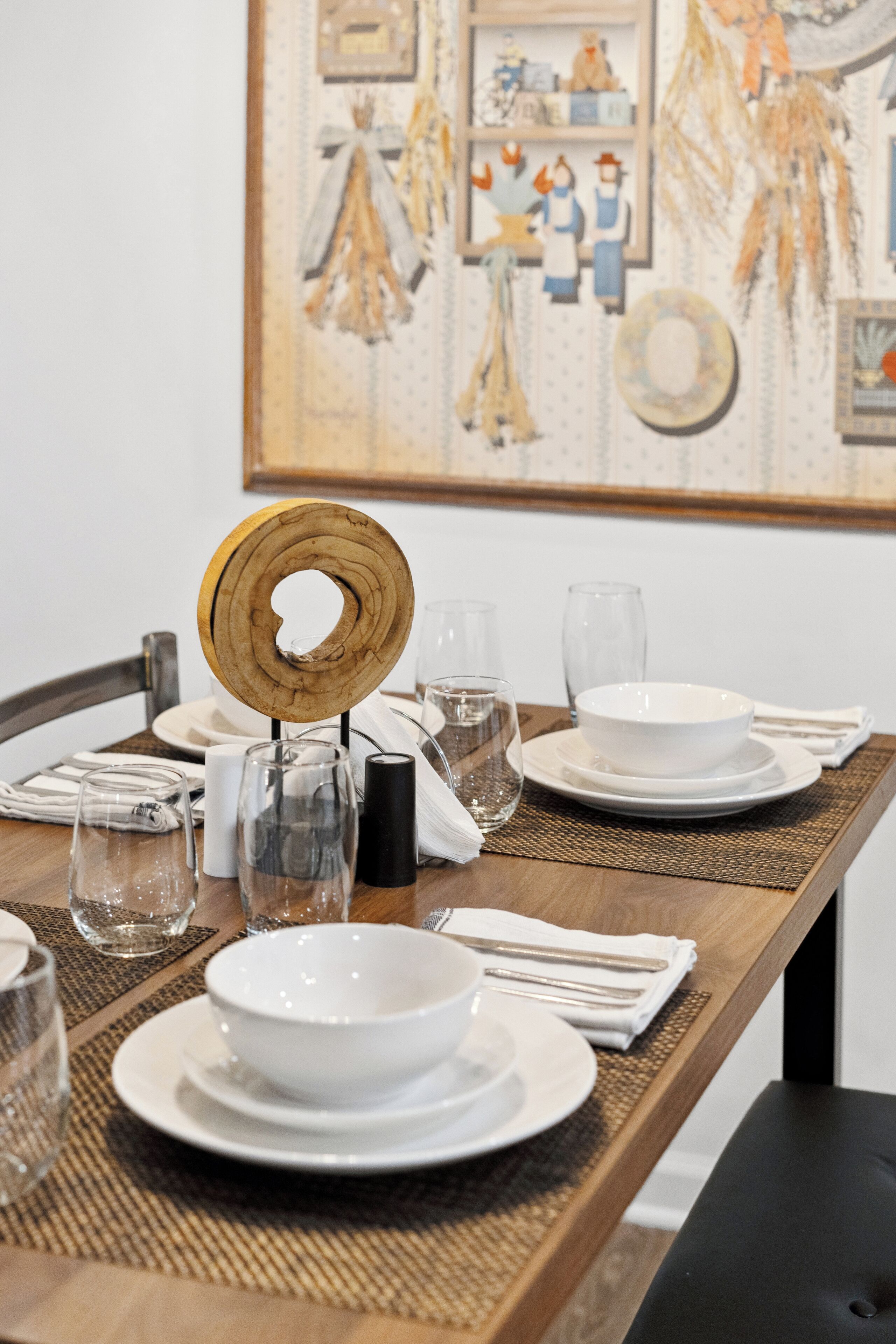 A dining table set with white plates, bowls, and clear glasses, with a wooden centerpiece and a framed artwork on the wall behind.