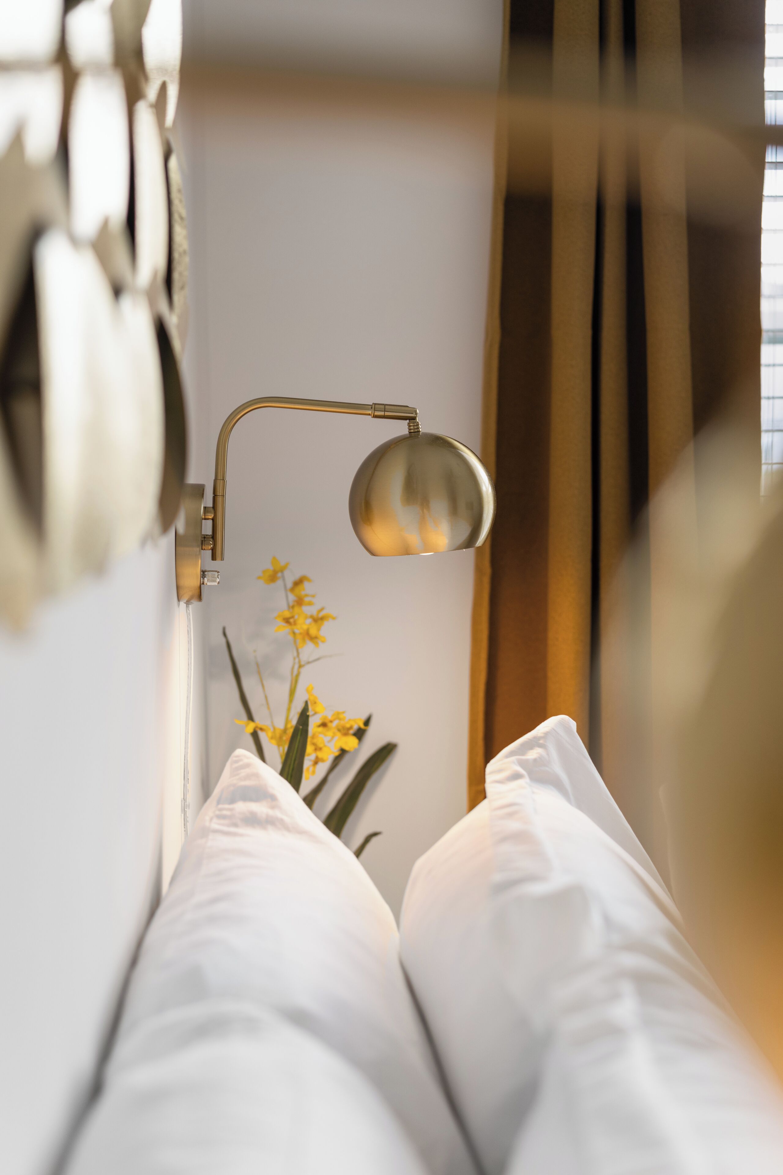 A bed with white bedding, a wall-mounted lamp, and a potted plant with yellow flowers.