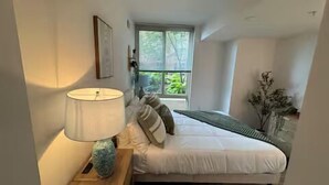 Ein Schlafzimmer mit Bett, Nachttischlampe, einem Fenster mit Blick auf Grünfläche und einem Wandbild.