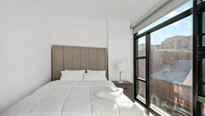 A hotel room with a large bed, a nightstand, a lamp, and a window with a city view. - Shaw #501 | 2BR | Balcony, City Views & Garage (washington)