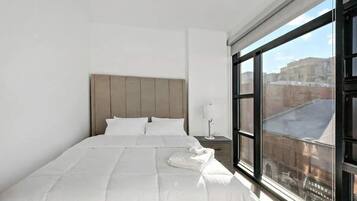 Ein Hotelzimmer mit einem großen Bett, einem Nachttisch, einer Lampe und einem Fenster mit Blick auf die Stadt.