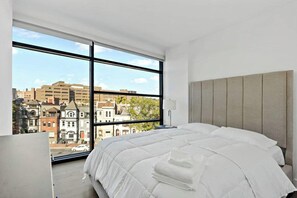 A hotel room with a large window, a bed with white bedding, and a view of buildings outside. - Shaw #501 | 2BR | Balcony, City Views & Garage (washington)