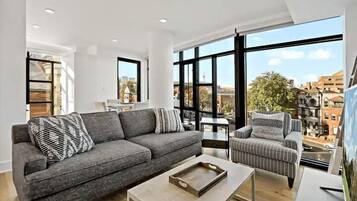 A modern living room with a grey sofa, striped armchair, and a coffee table. Large windows offer a view of the city.