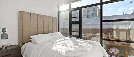 A modern bedroom with a large bed, a nightstand, and a window with a view of buildings.