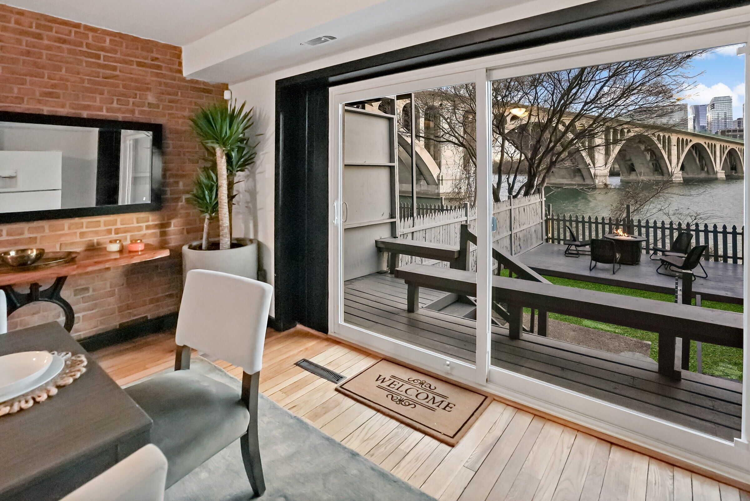 A modern interior with a dining area, a large sliding glass door leading to an outdoor deck with a view of a bridge and river.