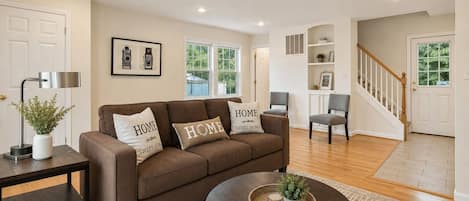 A living room with a brown sofa, two pillows with 'HOME SWEET HOME' text, a small table with a plant, and a staircase leading to another room.