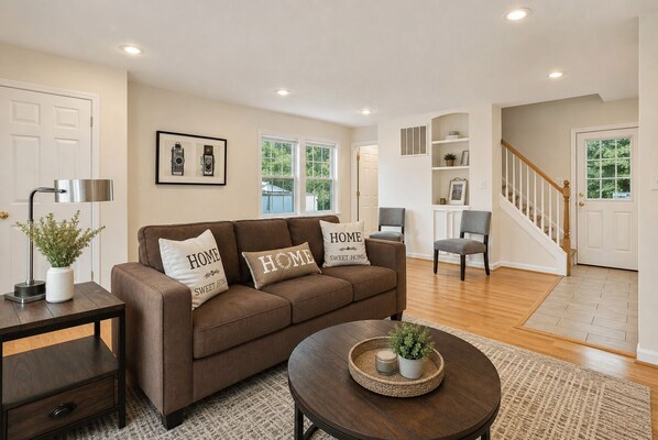 A living room with a brown sofa, two pillows with 'HOME SWEET HOME' text, a small table with a plant, and a staircase leading to another room.