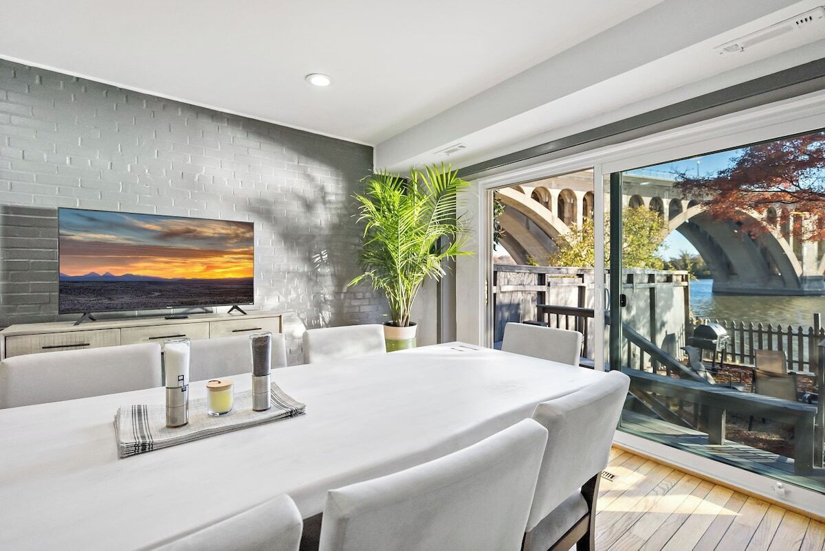 A modern dining area with a large table, white chairs, a TV on the wall, and a view of a bridge and water.