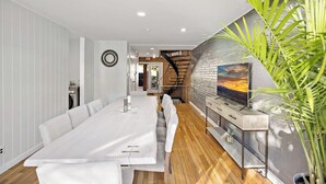Ein moderner Essbereich mit einem langen weißen Tisch, einer Couch und einem an einer Ziegelwand befestigten Fernseher.
