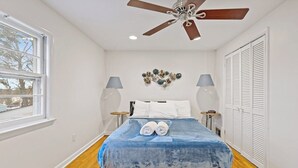 Un dormitorio con cama, ventilador de techo, dos lámparas de noche, una ventana y un adorno en la pared.