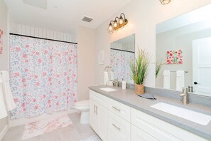 A bathroom with a floral shower curtain, a white toilet, a sink with a mirror, and a potted plant. - The Audrey The Yard District Downtown (Newport News)