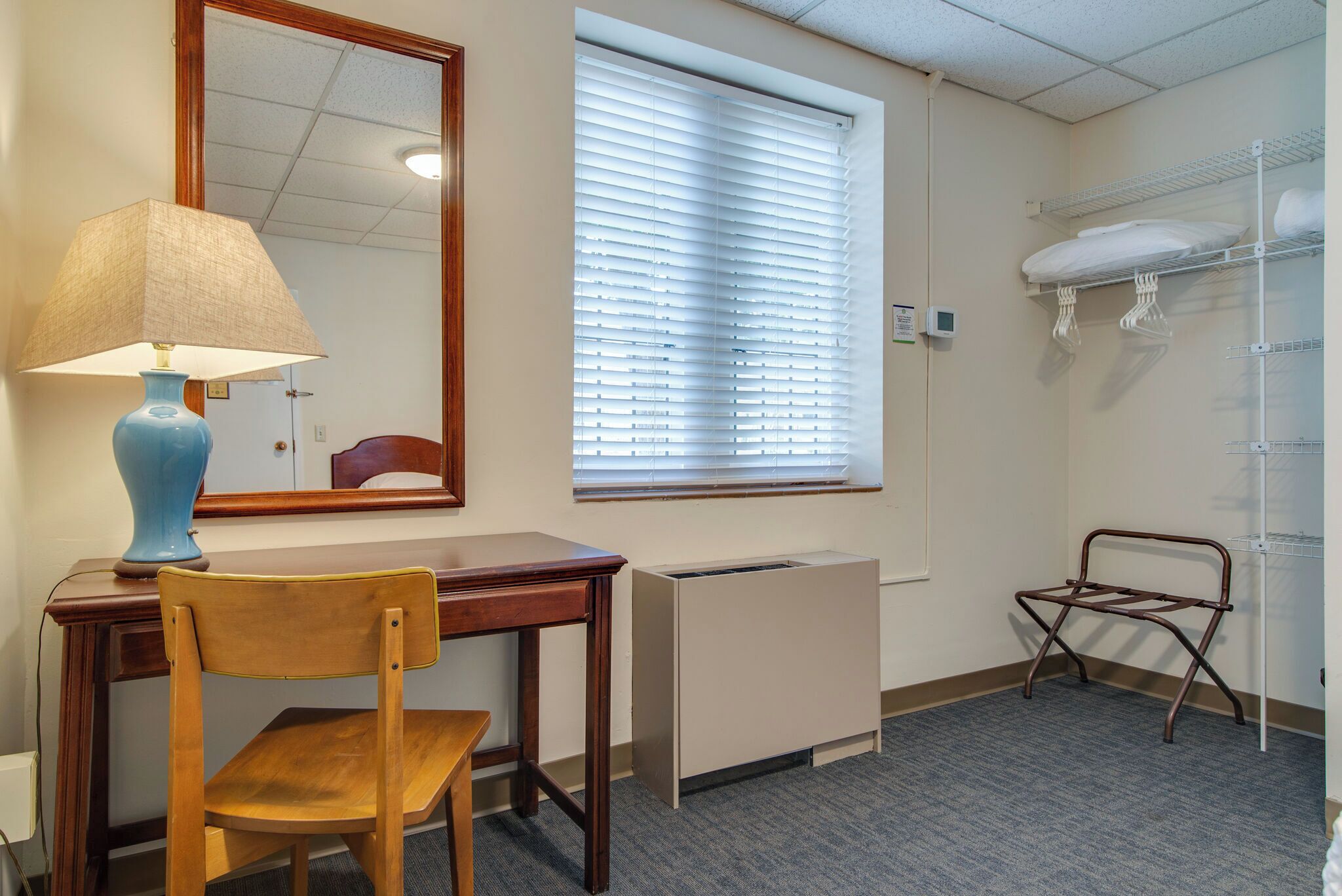 Une chambre d’hôtel avec un bureau en bois, une chaise, une lampe, un miroir, une fenêtre avec des stores, un radiateur et une étagère en métal avec des serviettes.