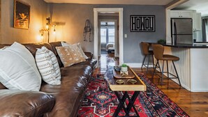 A living room with a brown leather sofa, patterned cushions, a red and black rug, and a wooden coffee table with decorative items.
