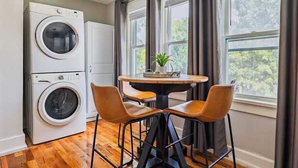A laundry room with a front-loading washer and dryer, a small table with chairs, and a window with curtains.