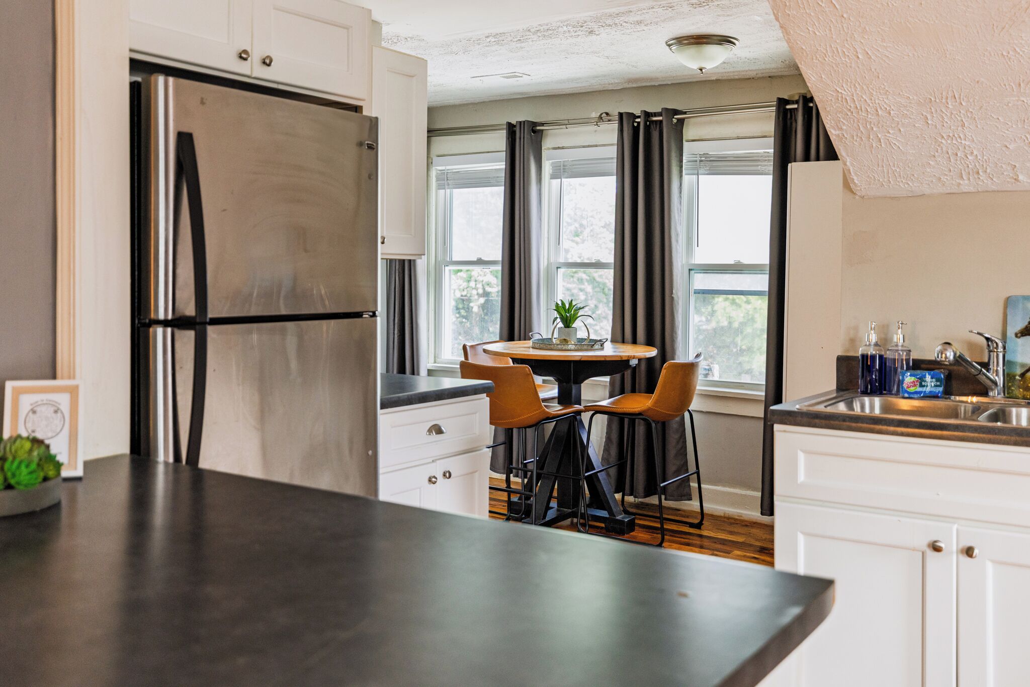 Eine moderne Küche mit einem Edelstahl-Kühlschrank, weißen Schränken, einer dunklen Arbeitsplatte und einem Essbereich mit zwei Stühlen und einem Tisch.