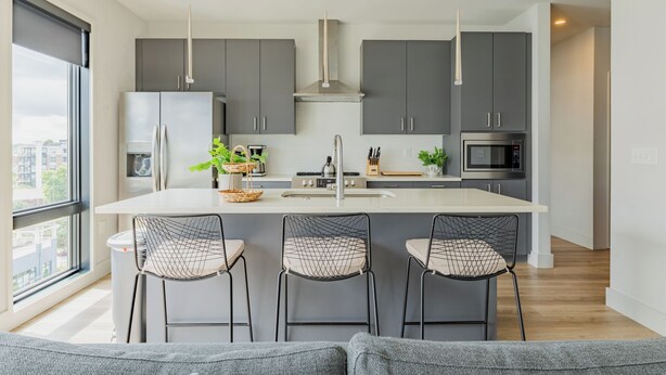 A modern kitchen with a central island, grey cabinets, and a large window. - Omnia - Modern Luxe unit at The Odyssey! (Nashville)