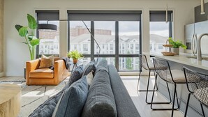 A modern living room with a sofa, a coffee table, and a dining area with chairs. Large windows provide a view of buildings outside. - Omnia - Modern Luxe unit at The Odyssey! (Nashville)