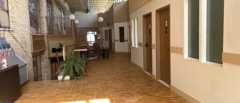 A spacious indoor corridor with a high ceiling, brick walls, and a tiled floor. There are doors numbered 2 and 3, a bench, and a potted plant.