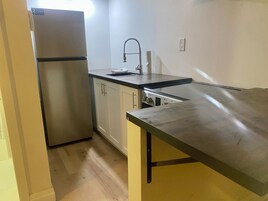 Una cocina compacta con un refrigerador de acero inoxidable, gabinetes blancos, una encimera negra y un lavamanos.