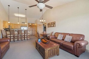 Een woonkamer met een bruine leren bank, een houten salontafel en een eetgedeelte met houten stoelen, met een keuken op de achtergrond.