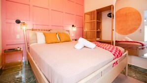 A bedroom with a pink wall, a bed with white and orange bedding, a wooden wardrobe, and a circular mirror.