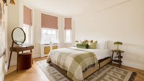 Une chambre à coucher comprenant un lit, une table de chevet, une lampe, une commode, un miroir et des fenêtres équipées de stores.