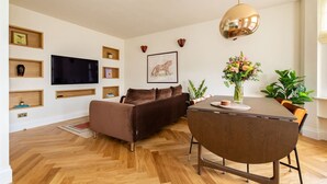 Un salon moderne avec un canapé marron, une table à manger avec des chaises, un téléviseur fixé au mur et une grande fenêtre.