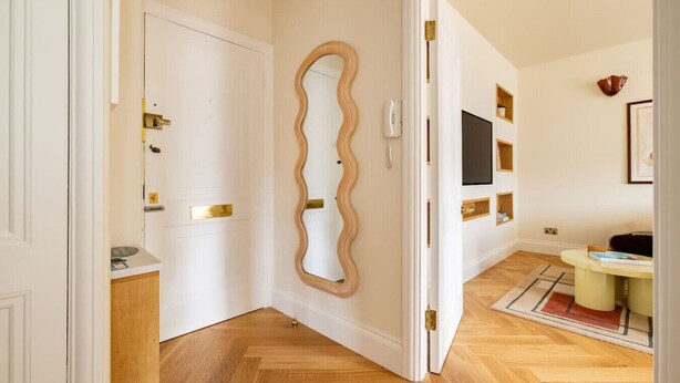 Un couloir doté d’un plancher en bois, d’une porte blanche avec des poignées en laiton et d’un miroir encadré d’un cadre ondulé.