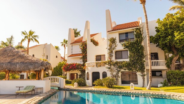 Un área de piscina con una estructura de techo de paja, una piscina y un edificio con balcones y un tejado de tejas rojas.