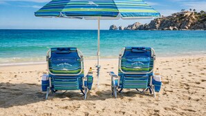 Dos sillas de playa con sombrillas y bolsas a juego sobre una playa arenosa, con vista despejada al océano y acantilados rocosos al fondo.