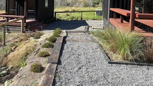 Un bâtiment avec un chemin de gravier menant à une terrasse en bois, entouré de végétation naturelle et situé au pied d’une colline.