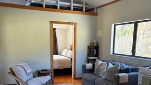 Un salon moderne avec un plancher en bois, un canapé gris, une chaise en bois, une petite table et un lit à l’arrière-plan.