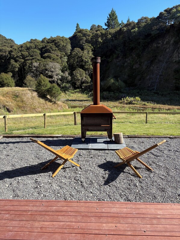 A rustic outdoor grill with two wooden folding chairs in a gravel area, surrounded by a wooden deck and a fenced grassy area with trees in the background.