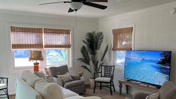 A living room with a sofa, armchairs, a television, a ceiling fan, and a large window with blinds.
